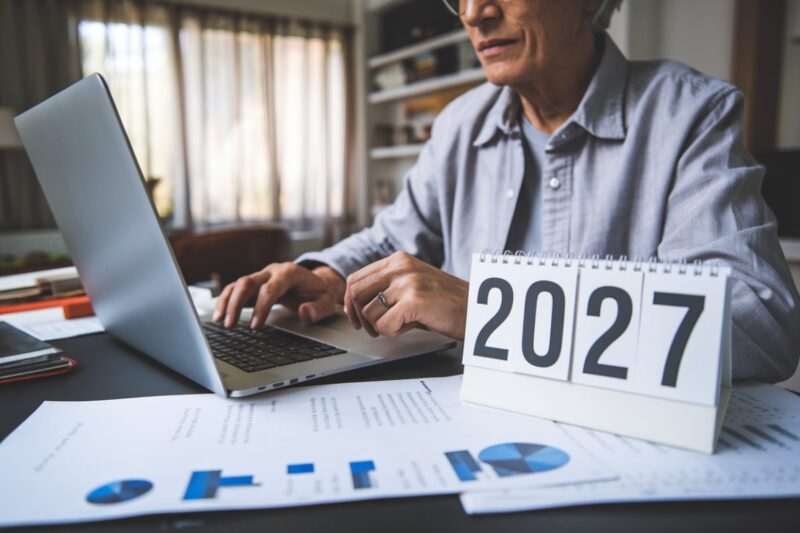 Personne âgée travaillant à domicile avec des documents financiers et un calendrier marqué 2027.